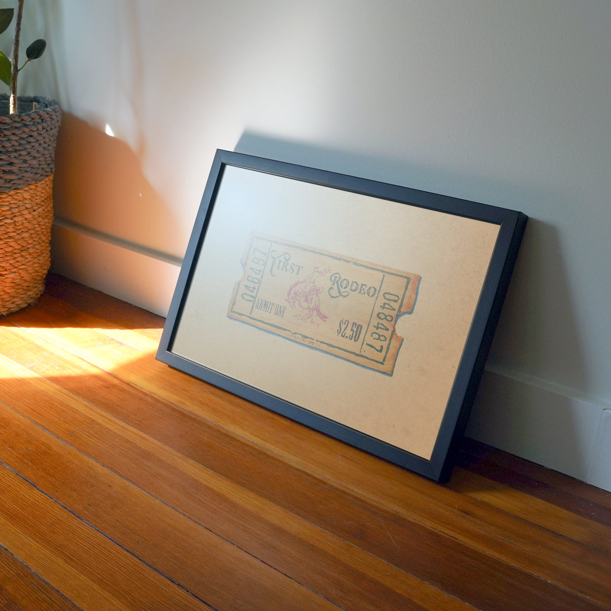 A framed art print featuring a vintage illustrated ticket on a cream background, set in a black frame and leaning against a white wall on a wooden floor.