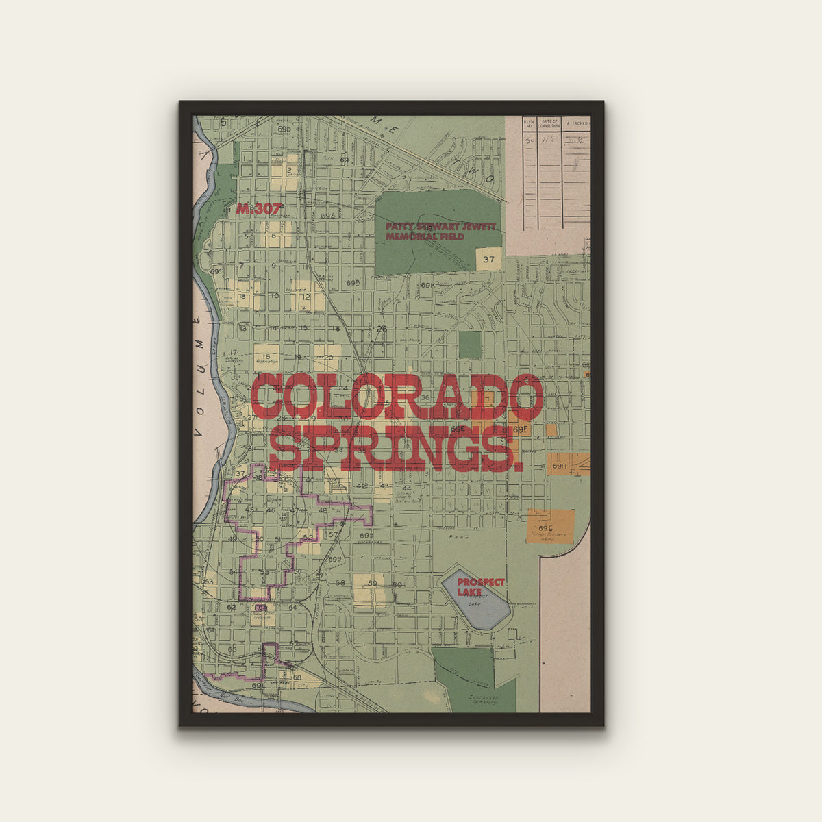 A framed vintage-style map print of Colorado Springs, Colorado, featuring a muted green street grid, red 'COLORADO SPRINGS' lettering, and a black frame against a light beige wall.