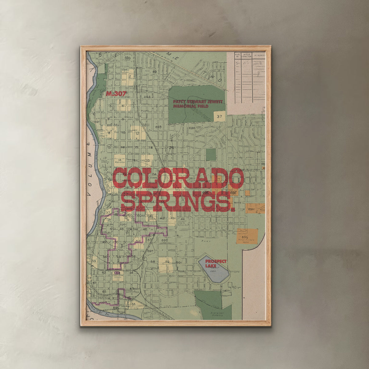 A framed vintage-style map print of Colorado Springs, Colorado, featuring a light wood frame and bold red text reading 'COLORADO SPRINGS' across the center, displayed against a textured gray wall.