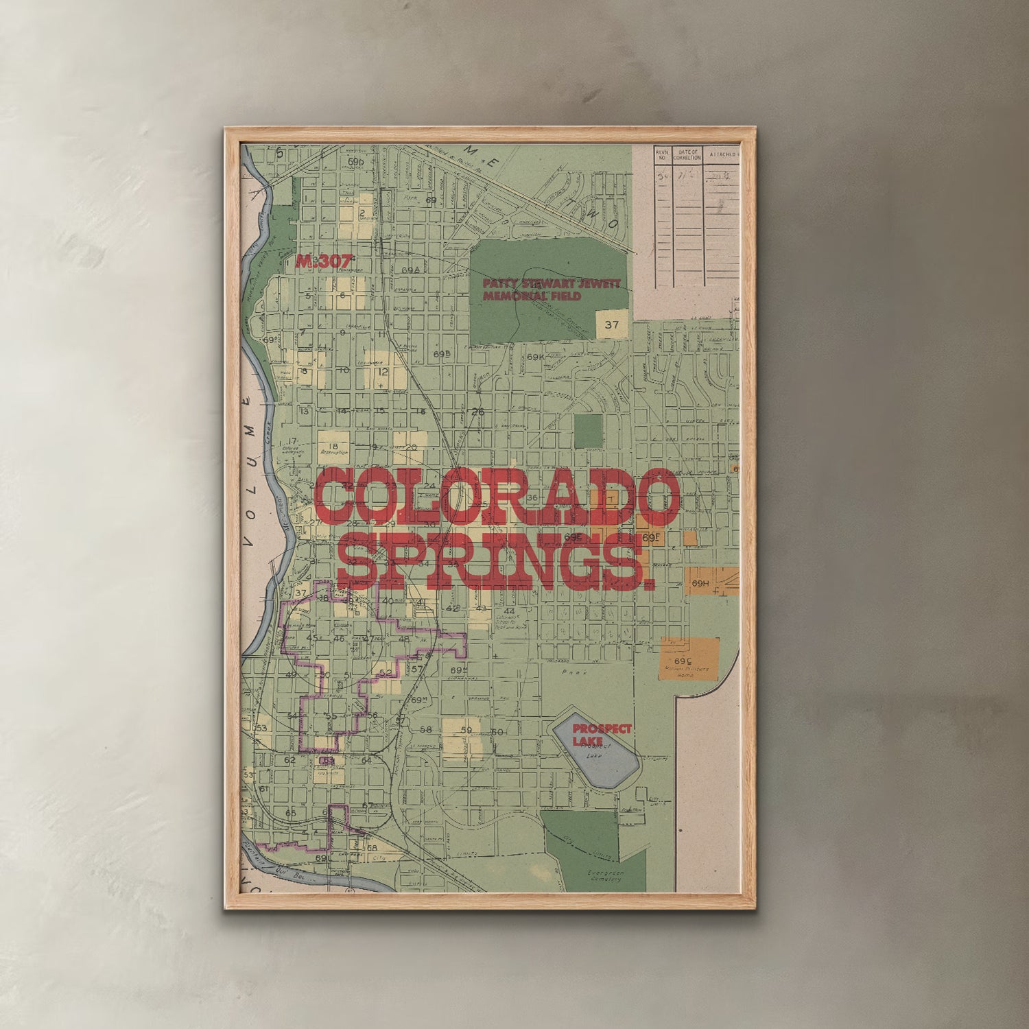 A framed vintage-style map print of Colorado Springs, Colorado, featuring a light wood frame and bold red text reading 'COLORADO SPRINGS' across the center, displayed against a textured gray wall.