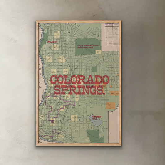 A framed vintage-style map print of Colorado Springs, Colorado, featuring a light wood frame and bold red text reading 'COLORADO SPRINGS' across the center, displayed against a textured gray wall.