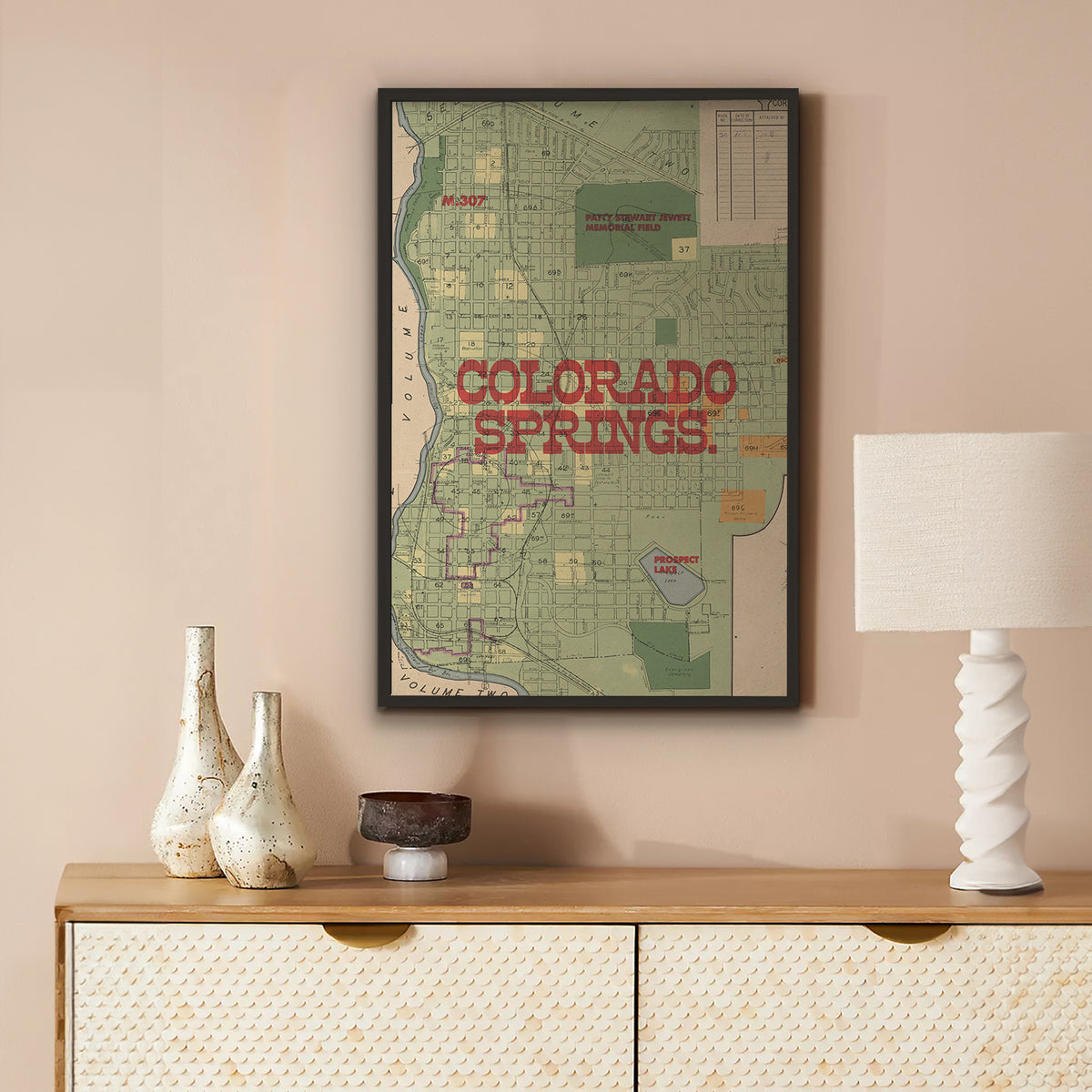 A framed vintage-style Colorado Springs map print hangs on a beige wall above a wooden console table with decorative vases, a bowl, and a white lamp.