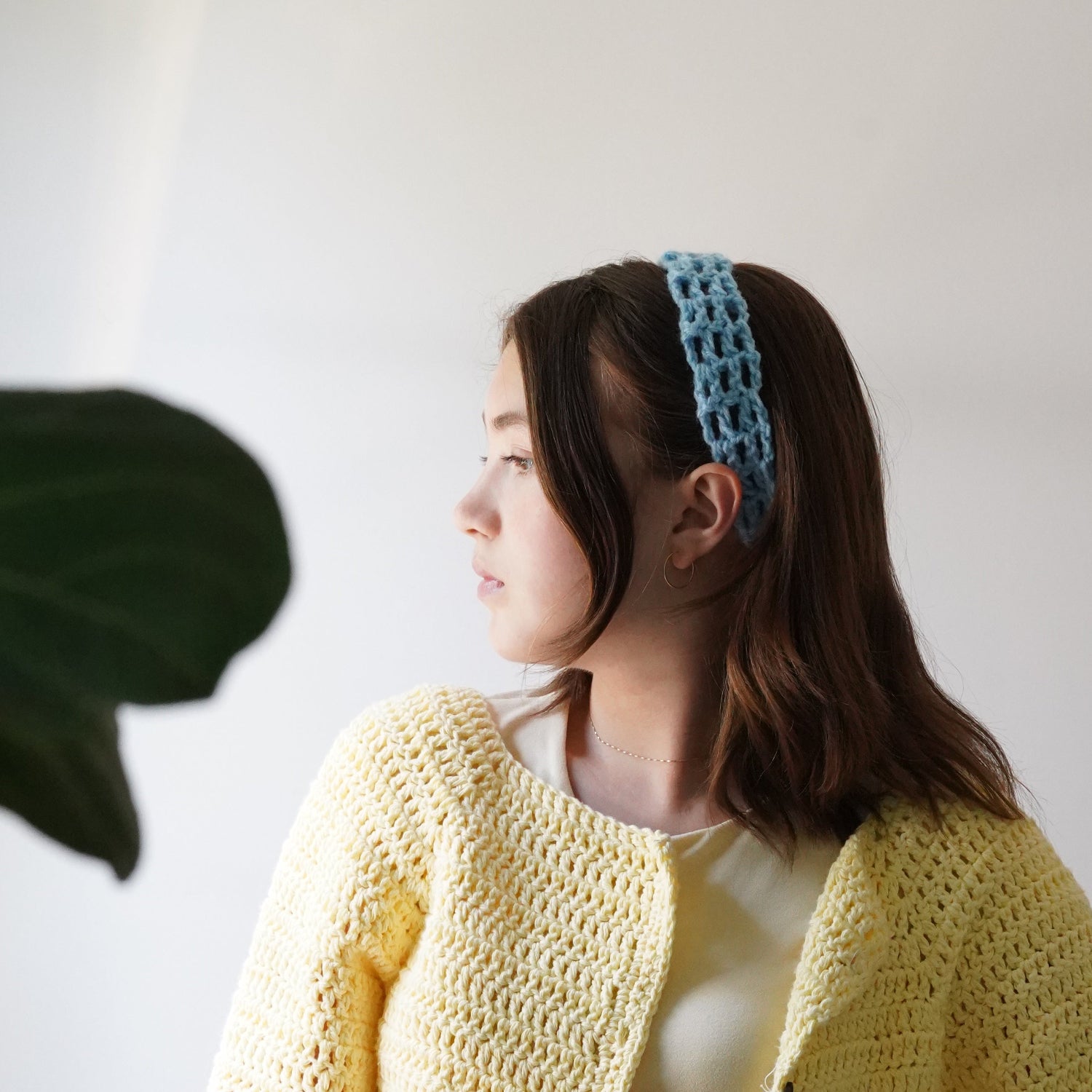 Woman wearing a yellow crochet cardigan and blue crochet headband with a white wall and plant in the background