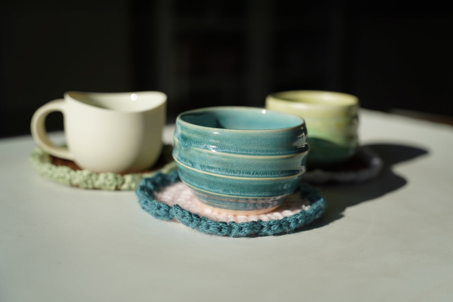 Three ceramic cups on crocheted coasters with a focus on a teal cup.