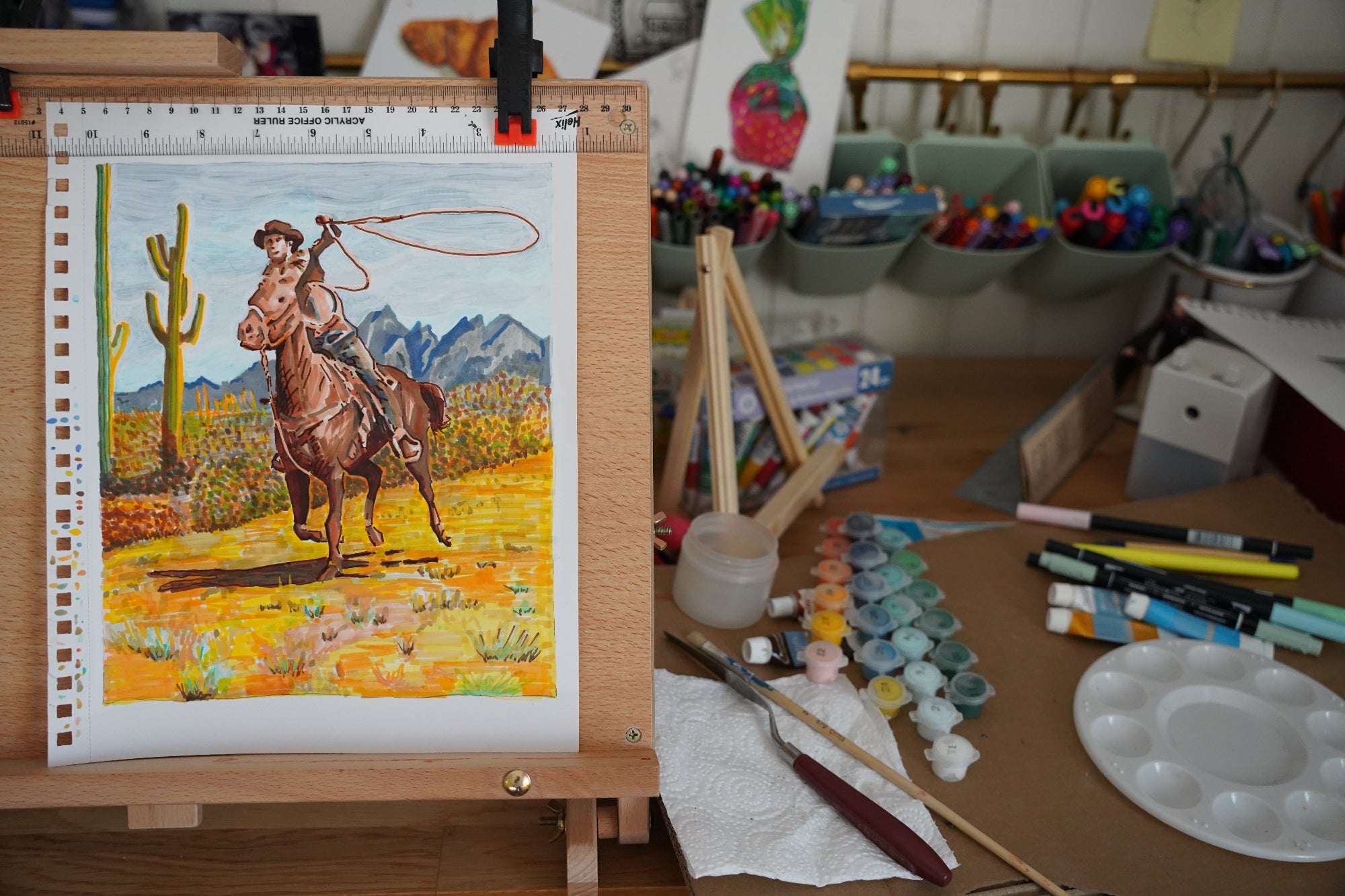 Painting of a cowboy on horseback with mountains in the background, displayed on an easel with art supplies around.